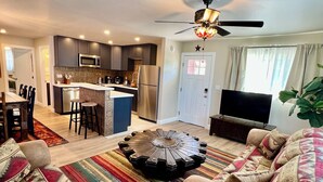A modern living room with a central coffee table, a sofa set, a television, and a kitchen area with a microwave, refrigerator, and bar stools.