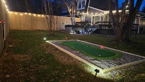 Un jardin avec un terrain de mini-golf, une clôture en bois et une maison illuminée.