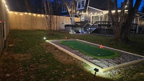 A backyard with a miniature golf course, a wooden fence, and a house with lights. - 1mi to Downtown |Hot Tub - Fenced Yard - Game Room (Waynesville)