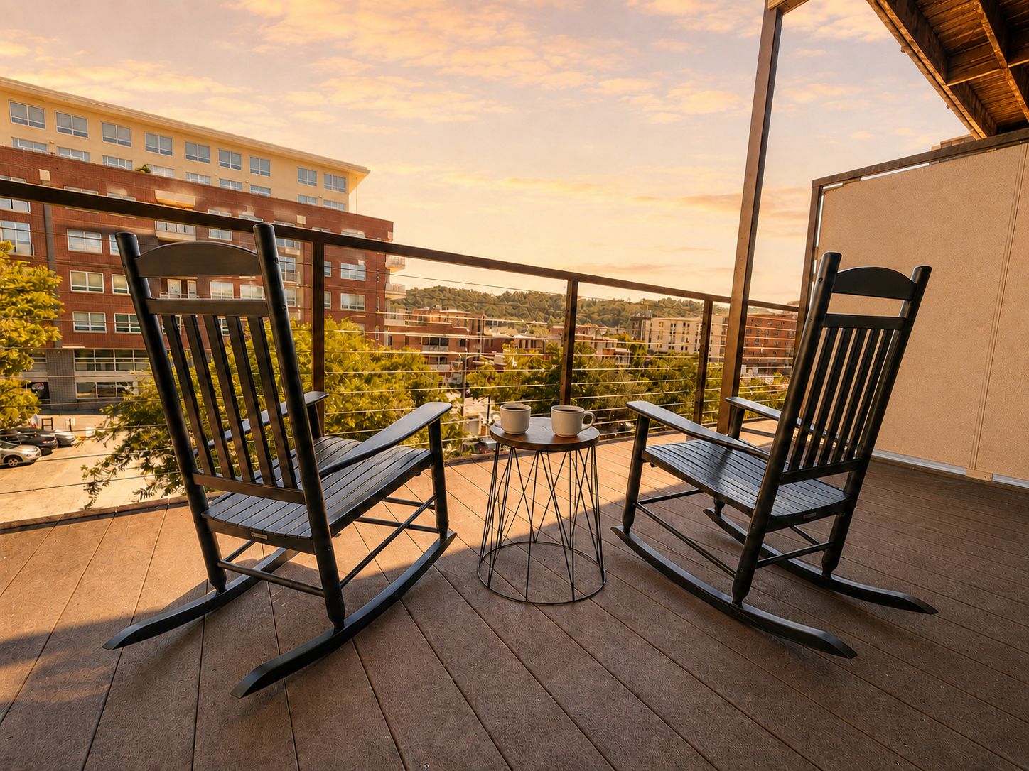 A balcony with two rocking chairs and a small table, overlooking a cityscape with buildings and trees.