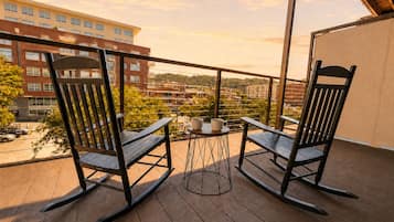 A balcony with two rocking chairs and a small table, overlooking a cityscape with buildings and trees.