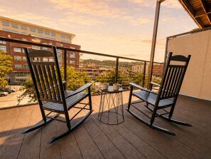 Un balcon avec deux fauteuils à bascule et une petite table, d’où l’on peut admirer un paysage urbain avec ses bâtiments et ses arbres.