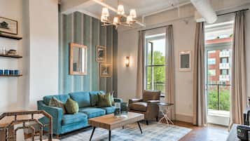 A living room with a blue sofa, a wooden coffee table, a leather armchair, and a patterned rug. There are framed pictures on the wall, a chandelier, and a window with curtains.