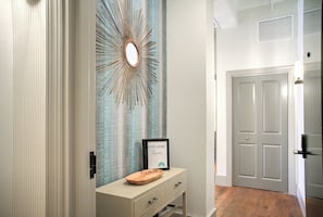 A hallway with a wooden floor, a white door, a framed welcome sign, and a decorative mirror.
