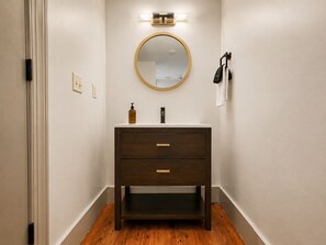 A bathroom with a dark wooden vanity, a round mirror, and a light fixture above the mirror. - The Gatsby at Heirloom Asheville | Downtown Asheville Historic Condo with Two King Suites (Asheville)