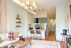 En moderne stue med sofa, sofabord og barstole. Et køkken med blå backsplash samt et spiseområde med bord og stole er synligt.