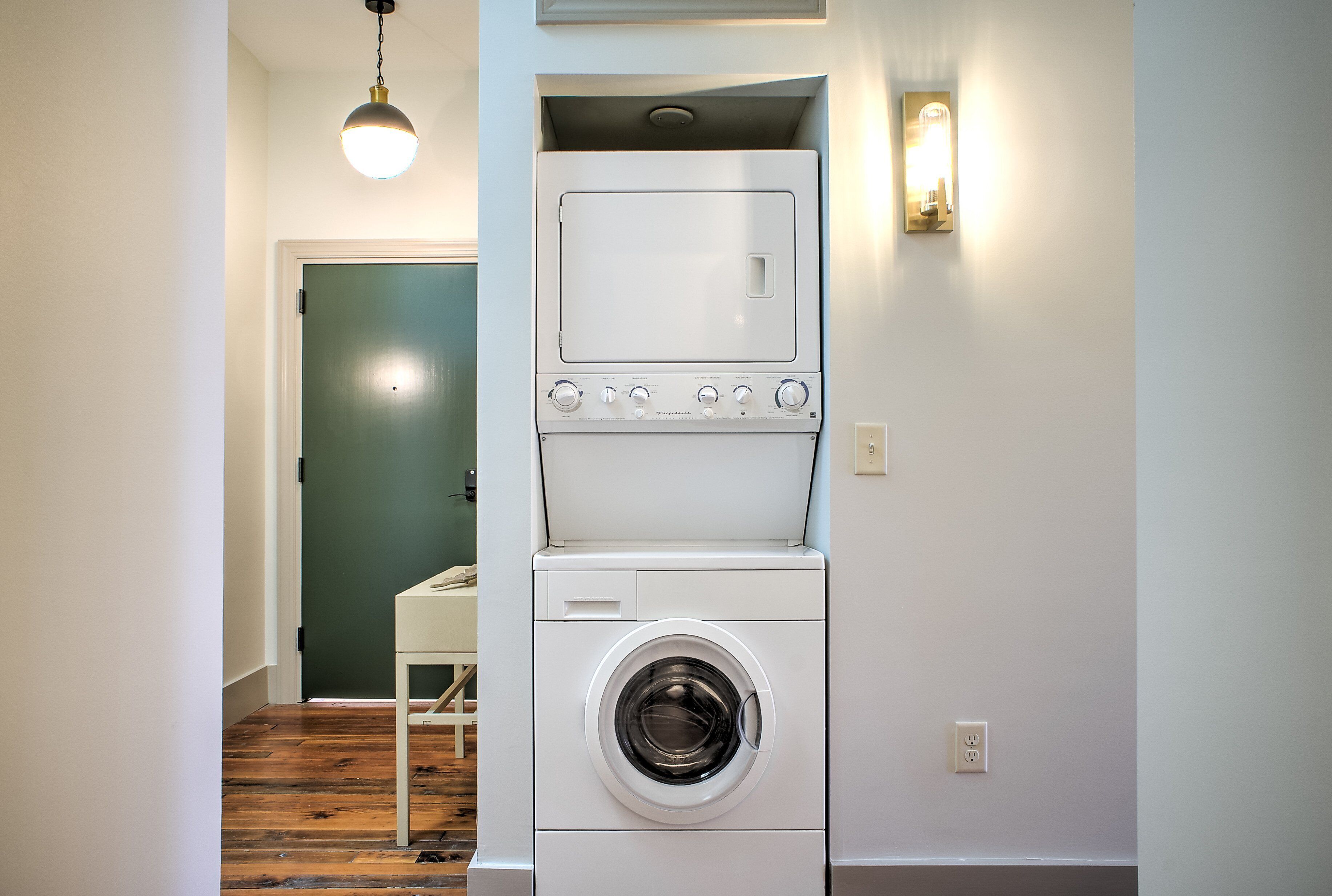 A compact laundry area with a front-loading washer and dryer, a wooden floor, and a green door in the background.