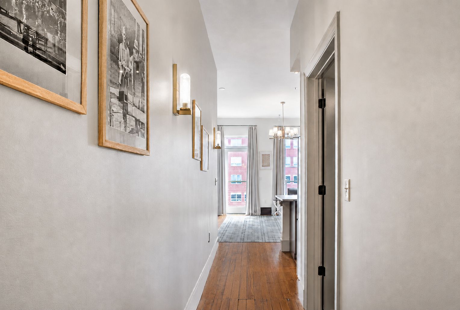 A hallway with framed pictures on the walls, a wooden floor, and a door leading to a room with a view of buildings outside.