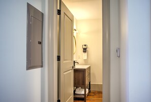 Un couloir étroit mène à une salle de bain équipée d’un meuble-lavabo en bois, d’un miroir et d’un porte-serviettes.
