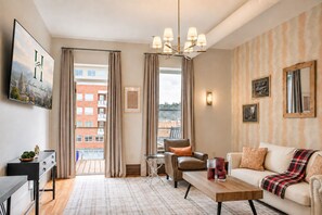 Un salon moderne avec un canapé, une table basse et un fauteuil. Il y a une grande fenêtre avec des rideaux, un lustre et un tableau au mur.