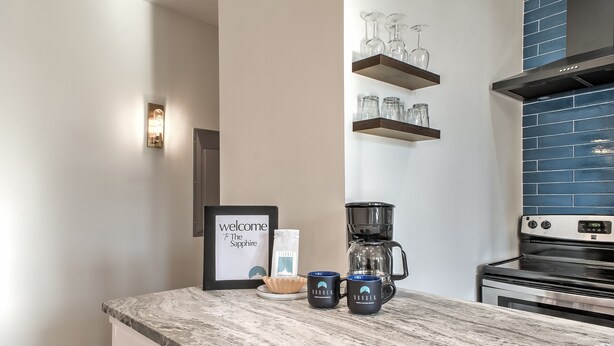 Eine Küche mit Marmorarbeitsplatte, einer Kaffeemaschine, zwei Tassen, einem gerahmten Willkommensschild und einem an der Wand montierten Regal mit Gläsern.