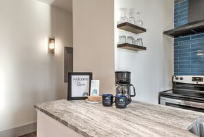 Eine Küche mit Marmorarbeitsplatte, einer Kaffeemaschine, zwei Tassen, einem gerahmten Willkommensschild und einem an der Wand montierten Regal mit Gläsern.