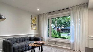 Ein modernes Wohnzimmer mit einem grauen Ecksofa, einem Holztisch und einem großen Fenster mit weißen Vorhängen.