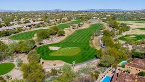 Ett flygbildsbild av en golfbana med flera greener, ett ökenlandskap och bostadshus i bakgrunden.