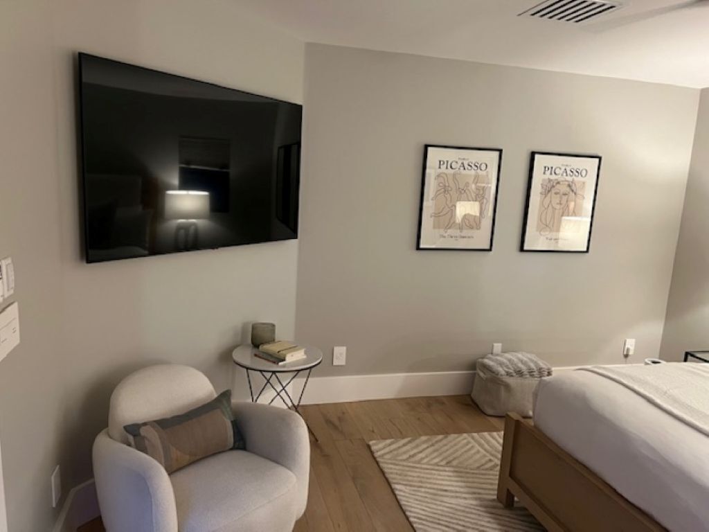 A modern bedroom with a large flat-screen TV, a bed, a chair, and two framed artworks on the wall.