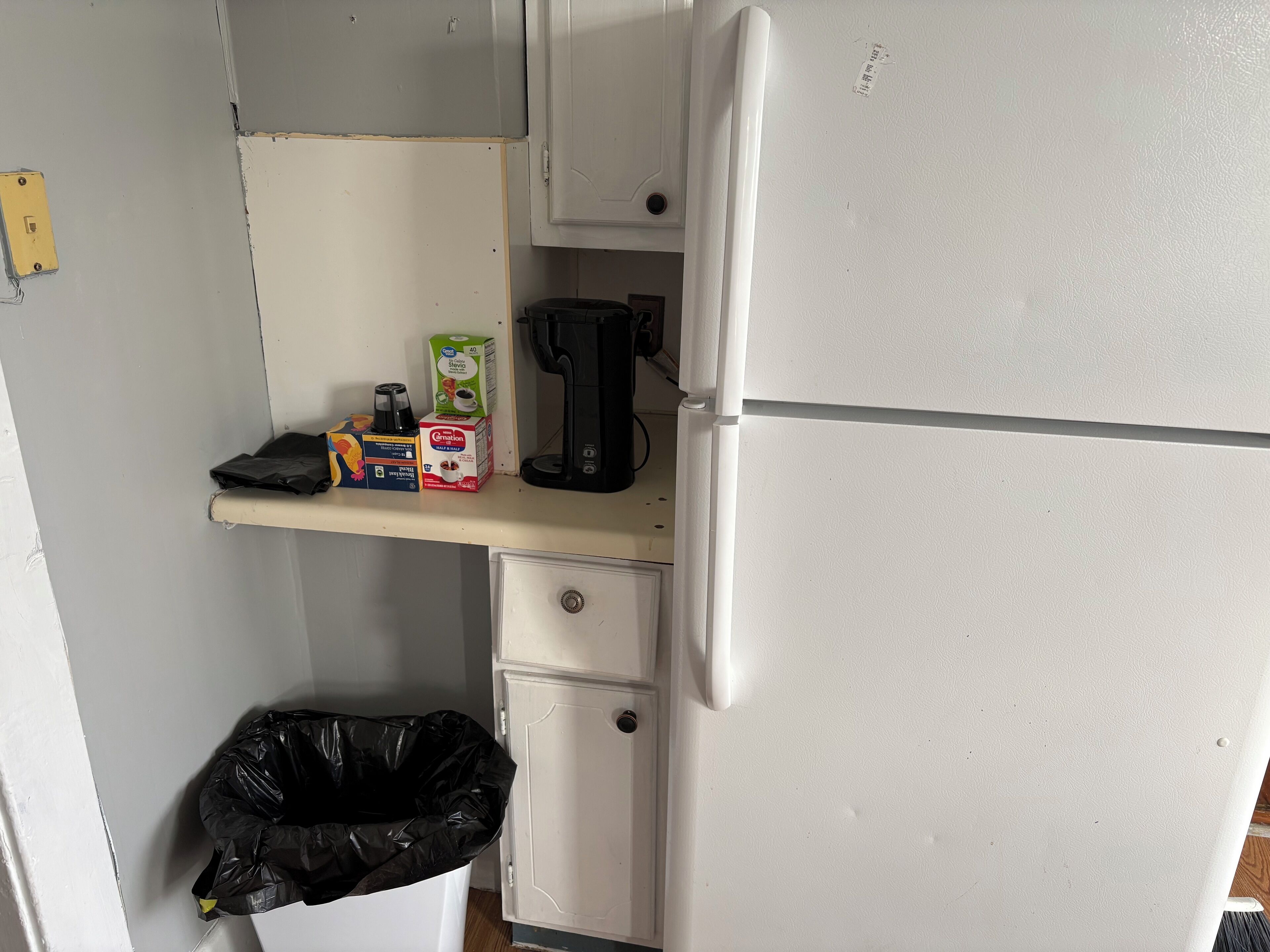 A kitchenette with a white refrigerator, a coffee maker, a box of cereal, and a trash bin.