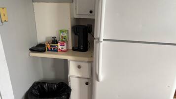 A kitchenette with a white refrigerator, a coffee maker, a box of cereal, and a trash bin.