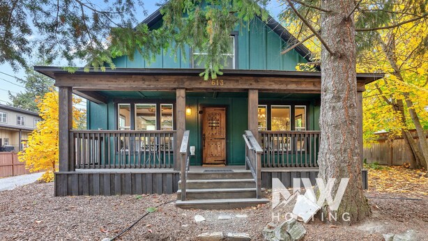 A two-story wooden cabin with a porch, numbered 613, surrounded by trees and a gravel driveway. - Downtown CDA Retreat – Hot Tub, Deck, & Walk to Lake! (Coeur D Alene)