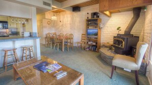 Een gezellige woonkamer met een open haard, een eethoek met stoelen, een keuken met een aanrecht en krukken, en een televisie op een houten dressoir.