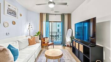 A modern living room with a white sofa, a round wooden coffee table, a ceiling fan, a flat-screen TV, and a balcony with a view of buildings.