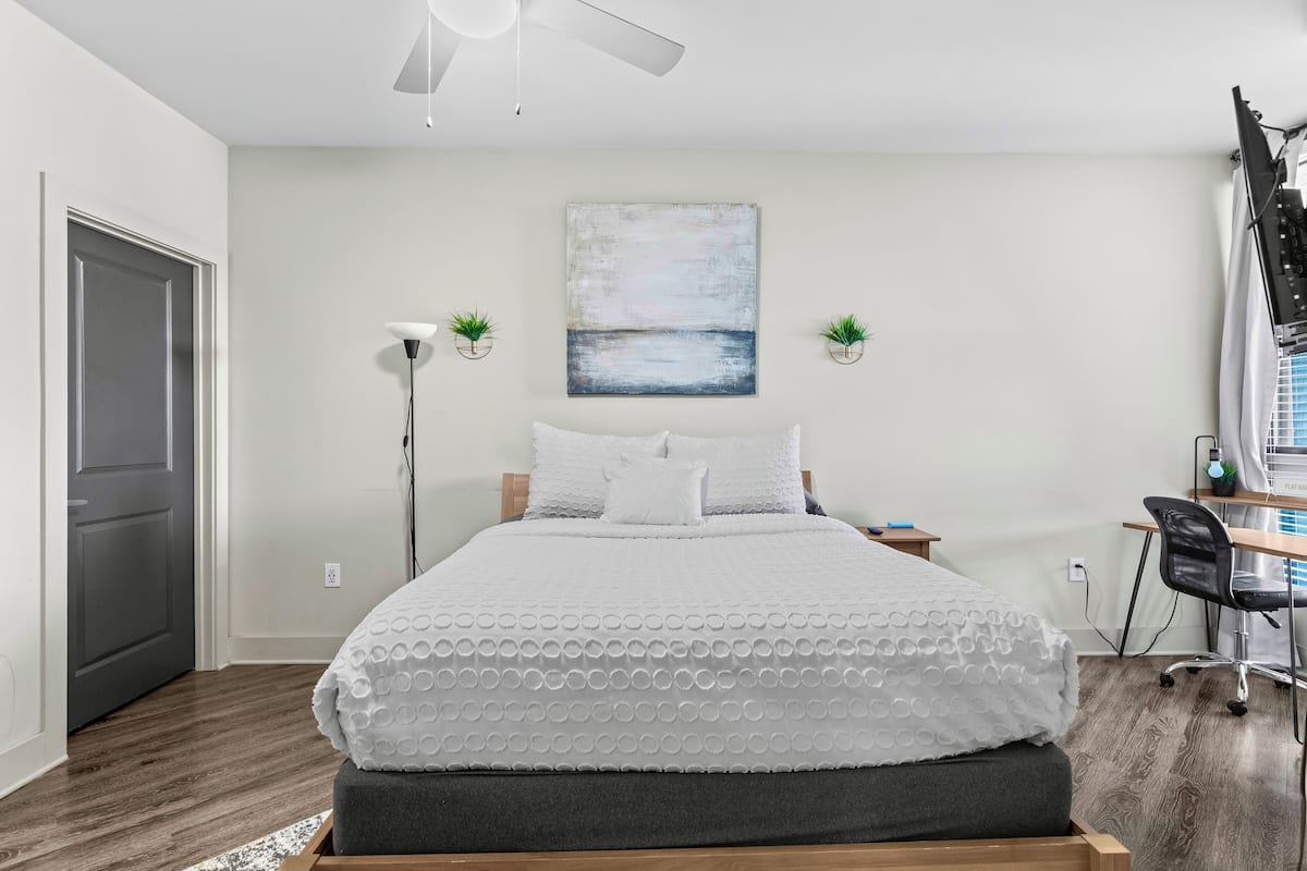 A bedroom with a bed, a desk, a chair, a ceiling fan, and a painting on the wall.