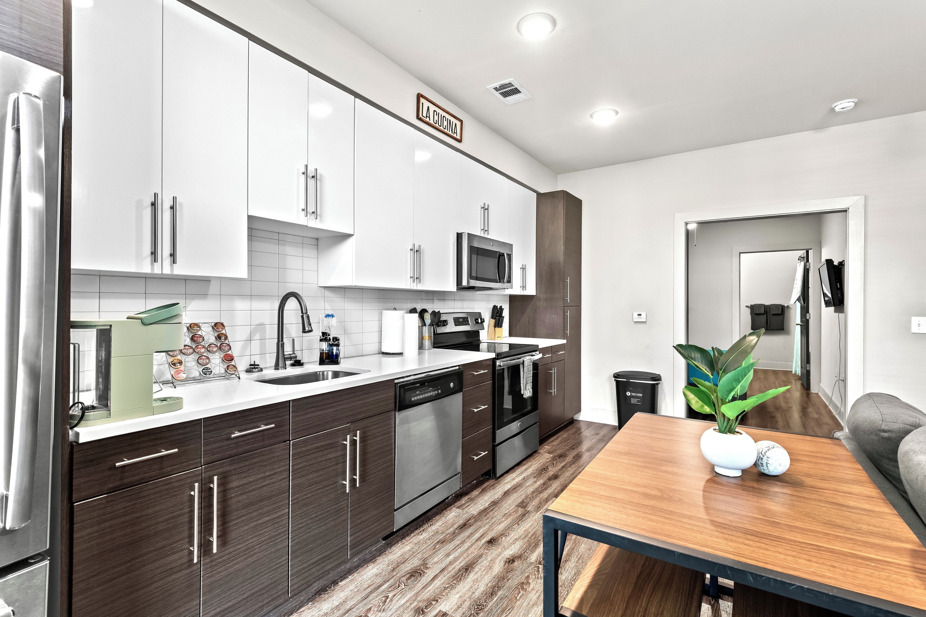 A modern kitchen with white and dark wood cabinets, stainless steel appliances, and a wooden dining table with a plant.