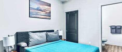 A neatly arranged bedroom with a bed, a nightstand, a wall-mounted lamp, a door, and a bathroom visible through an open door.