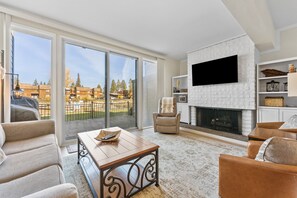 Ein geräumiges Wohnzimmer mit Kamin, großen Fenstern und einem Balkon mit Blick auf Gebäude und Bäume.