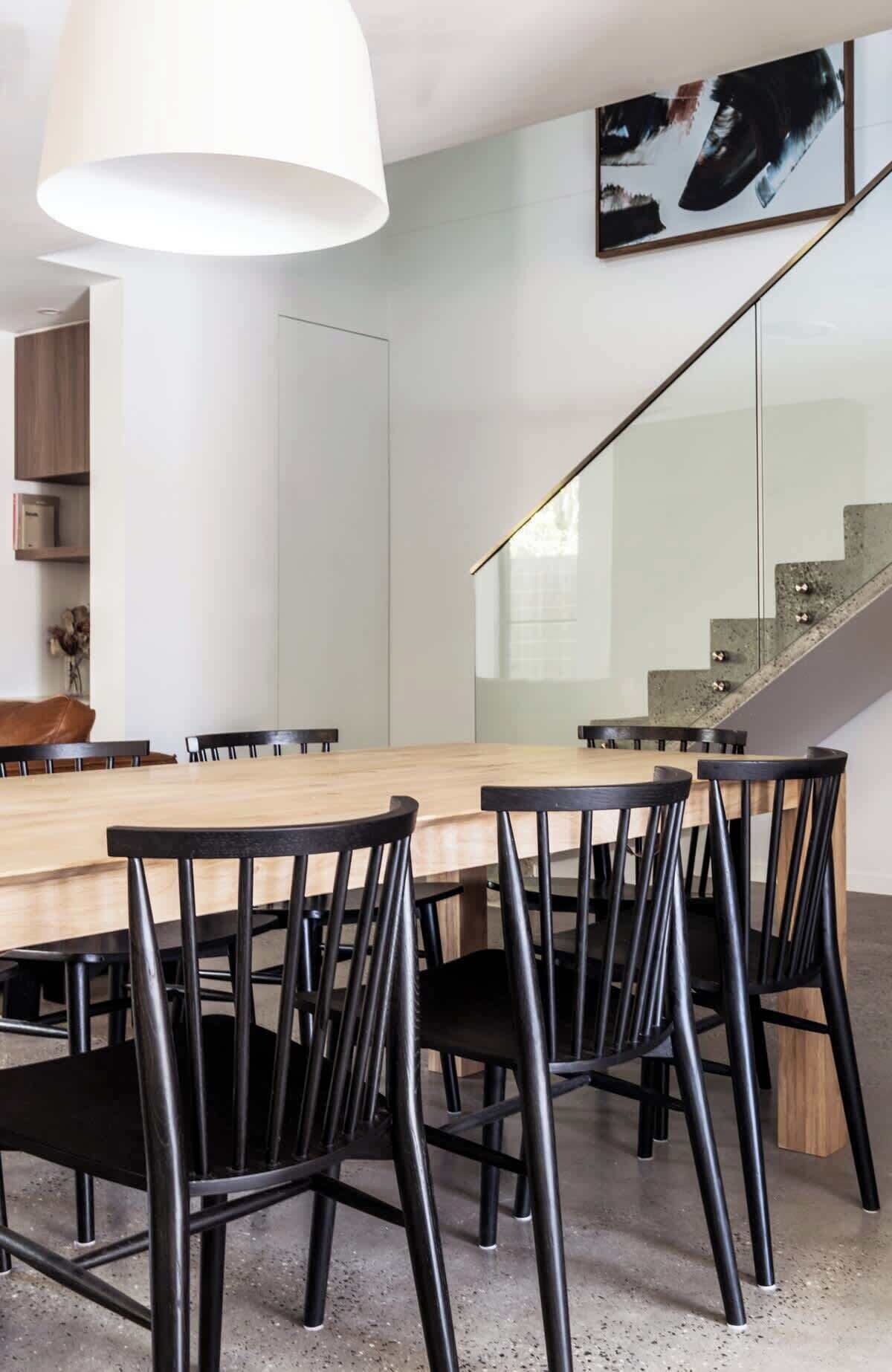 A modern dining area with a wooden table and black chairs, a white pendant light, and a staircase with a glass railing.