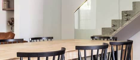 A modern dining area with a wooden table and black chairs, a white pendant light, and a staircase with a glass railing.