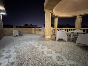 An outdoor patio area with a patterned floor, white plastic chairs, and a stone column structure. - Al Sawah 6BD Villa with private pool & garden @New Cairo (New Cairo)