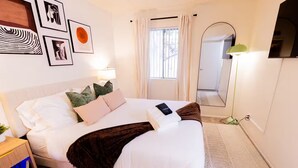 Un dormitorio ordenado con cama, almohadas, una mesita de noche, lámpara, espejo y cuadros en la pared.