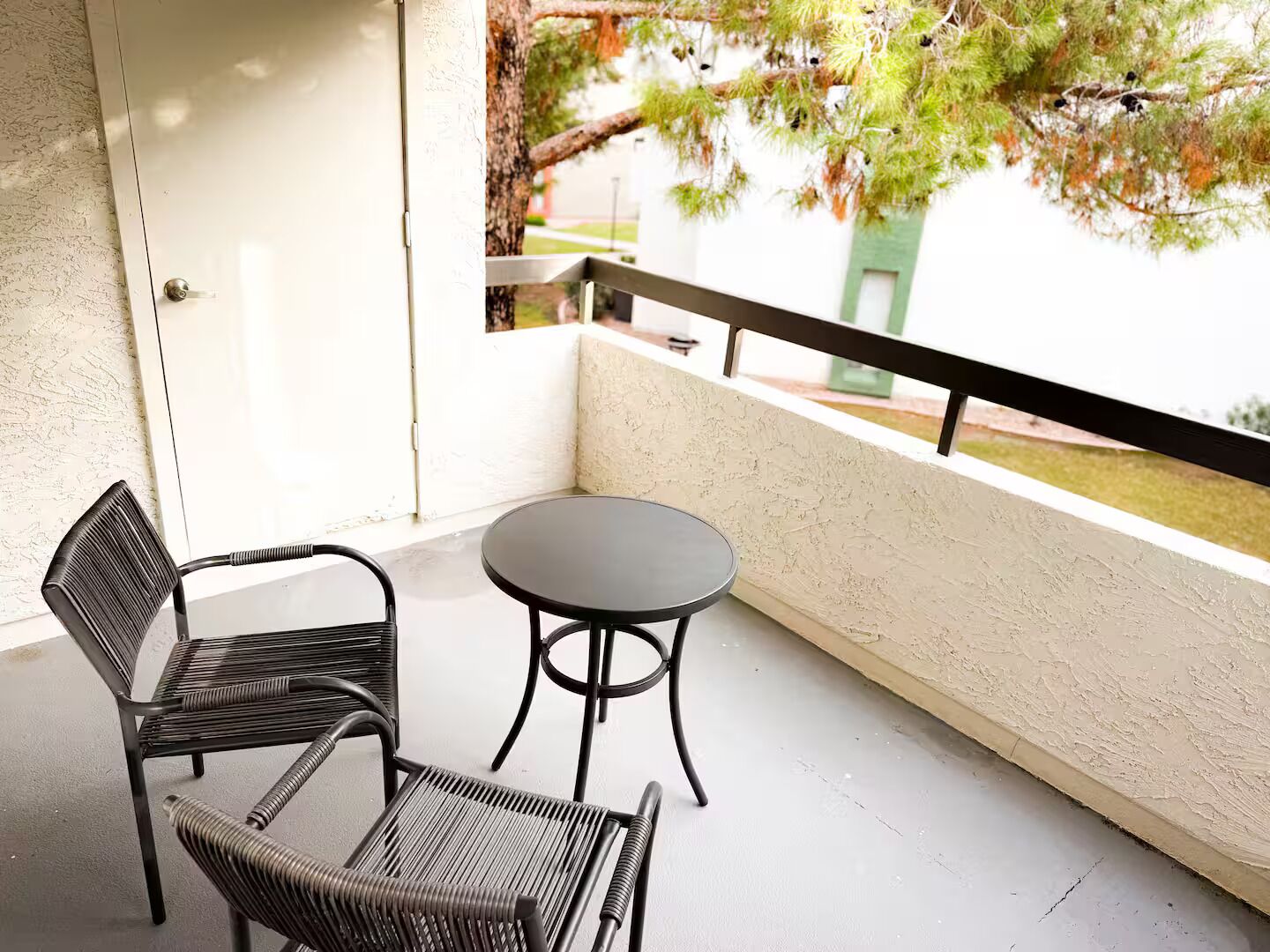 A balcony with a round table and two chairs, a white door, and a view of trees and a building.