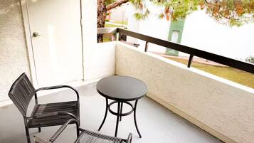 A balcony with a round table and two chairs, a white door, and a view of trees and a building.