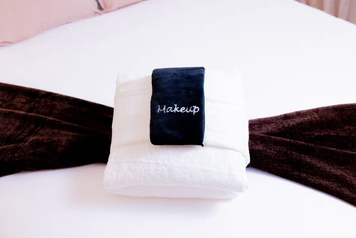 A neatly folded white towel with a black tag labeled 'Makeup' on a bed with a dark brown bedspread.