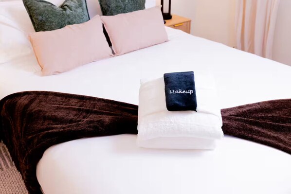A neatly made bed with a folded towel and a black pillow with "Makeup" embroidered on it.