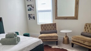 Een slaapkamer met een bed, een gestoffeerde fauteuil, een klein rond tafeltje en een grote spiegel.
