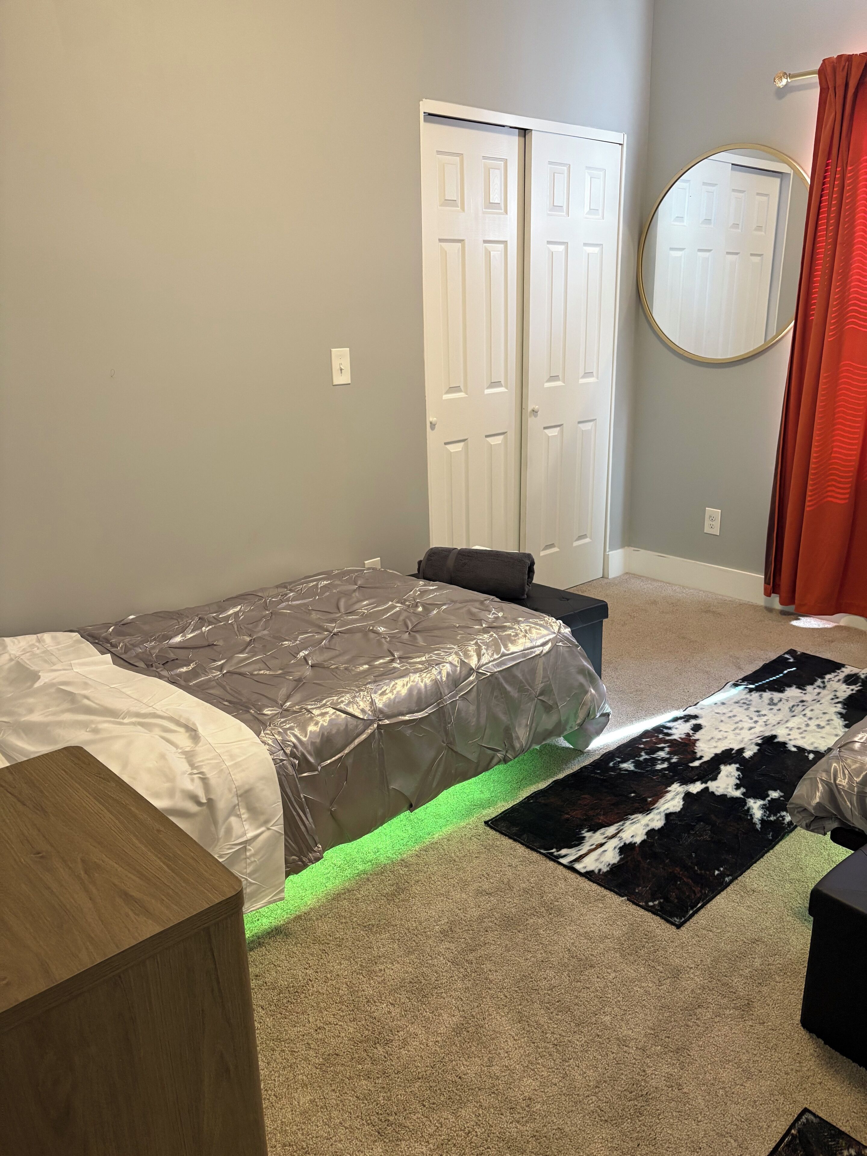 A bedroom with a bed, a wooden nightstand, a mirror, a door, and a rug.