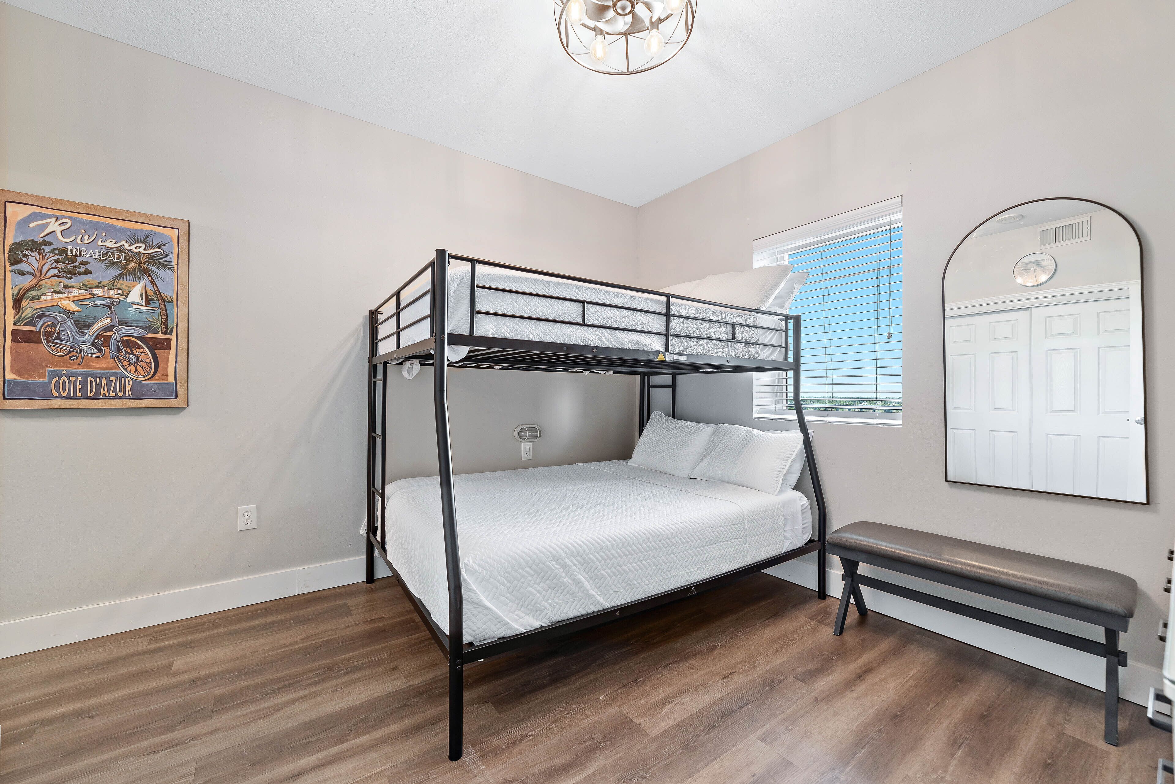 Una cama literera en una habitación con suelo de madera, un banco, una ventana con persianas y un espejo en la pared.