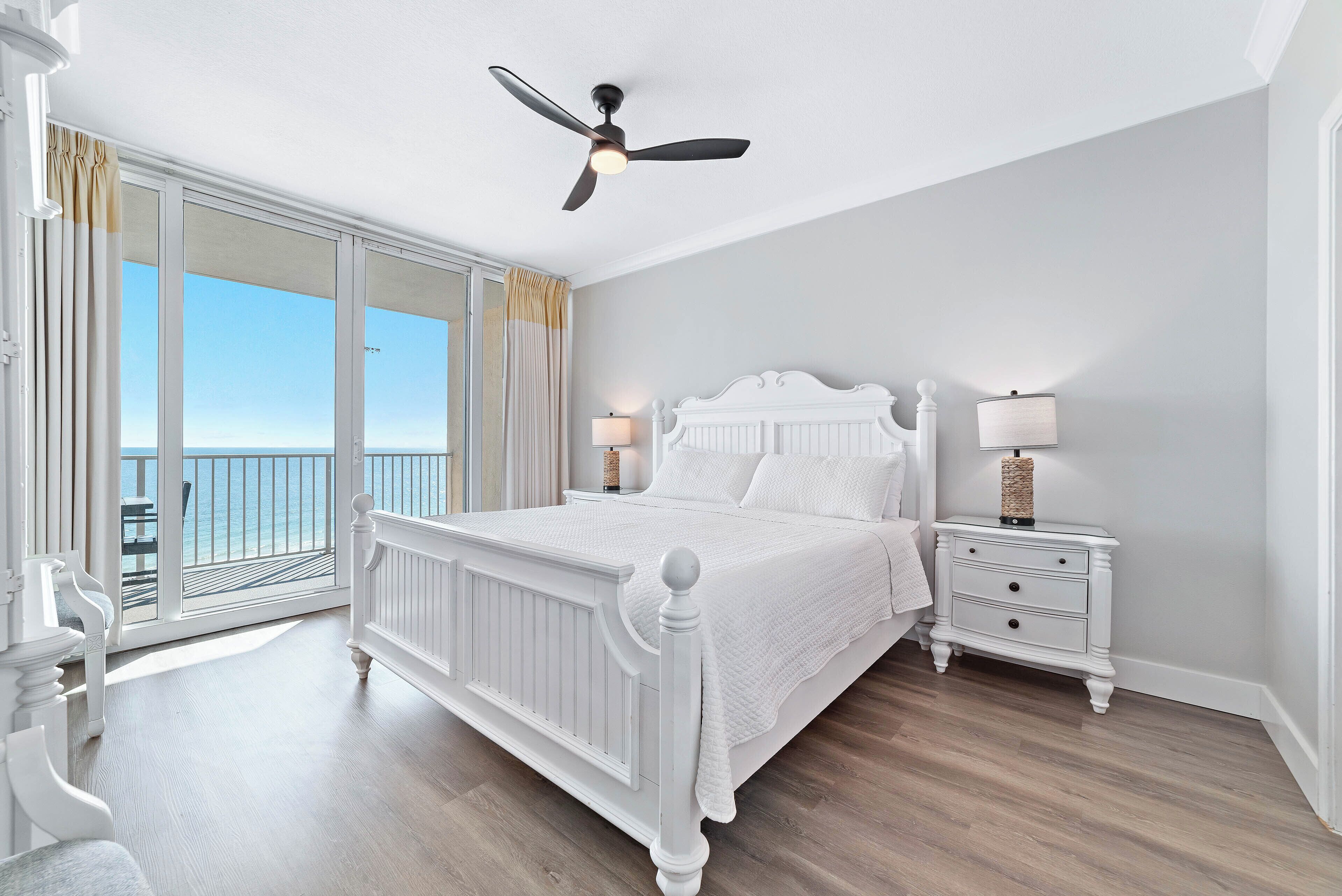 Quarto com cama, mesas de cabeceira, ventilador de teto e vista para o oceano através de amplas janelas.