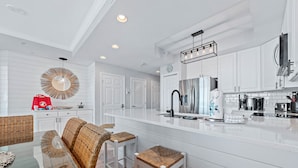 A modern kitchen with white cabinetry, a stainless steel refrigerator, and a breakfast bar with wicker stools. - Modern Beachfront Luxury | Balcony Grill, Gulf Views & Pool Access | BEACHBALL PROPERTIES (Gulf Shores)