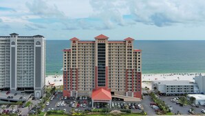 A high-rise hotel with a red roof, a beach view, and a parking lot with multiple cars. - Modern Beachfront Luxury | Balcony Grill, Gulf Views & Pool Access | BEACHBALL PROPERTIES (Gulf Shores)