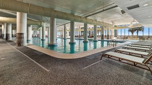 Ein Hallenbad mit mehreren Säulen, Liegestühlen und Blick auf Palmen im Freien.