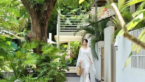 Une personne se promène dans un jardin luxuriant, où l’on trouve une grande variété de plantes tropicales, et un bâtiment blanc se dresse à l’arrière-plan.
