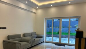 Un salon moderne avec un canapé d’angle gris, un sol carrelé brillant et une porte-fenêtre coulissante donnant sur une cour avec un petit jardin.