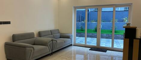 A modern living room with a grey sectional sofa, a glossy tiled floor, and a sliding glass door leading to a courtyard with a small garden.