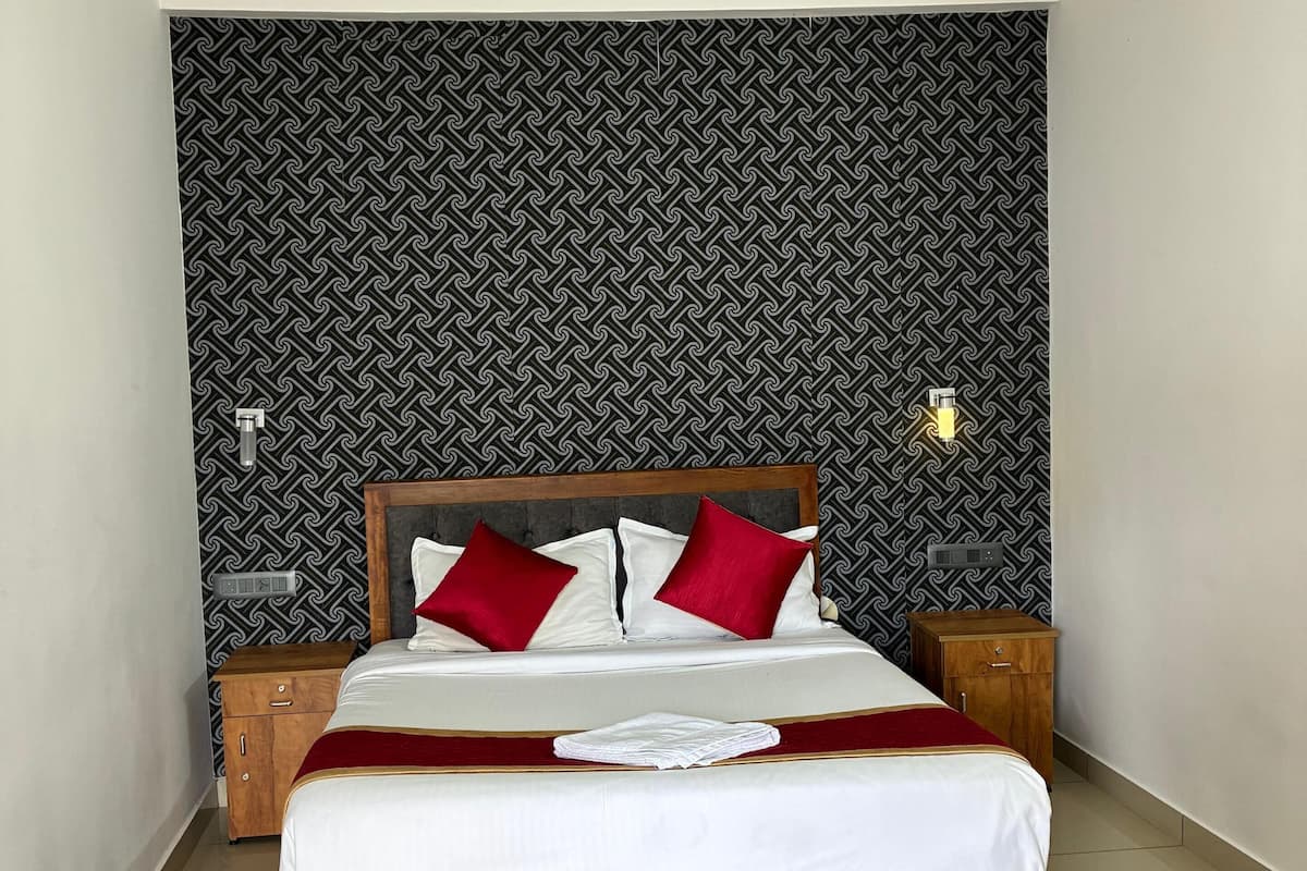 Una cama bien hecha con estructura de madera, dos almohadas rojas y una colcha blanca. Arriba, un ventilador de techo, y al lado, una mesita de noche de madera con una lámpara. La habitación tiene un papel tapiz estampado y piso de baldosas.
