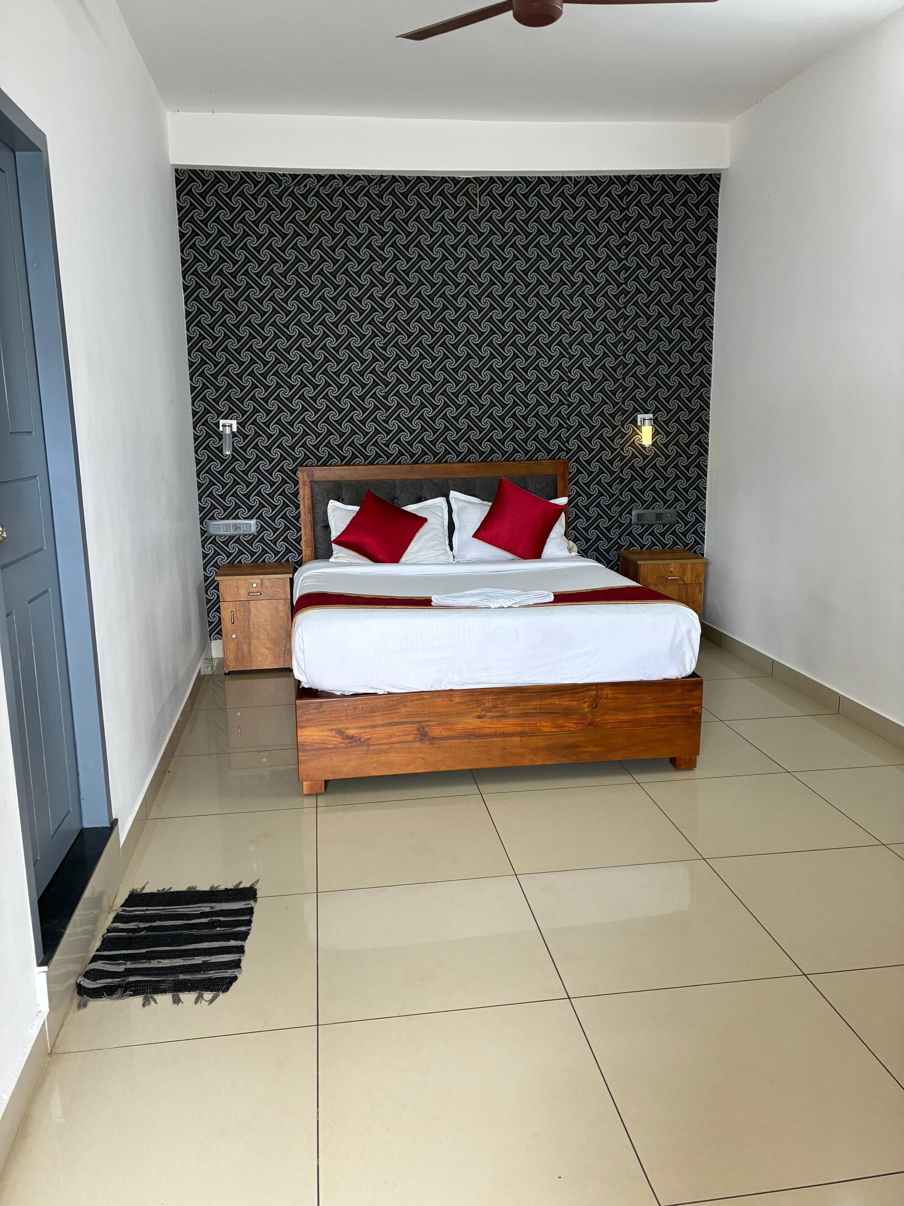 Quarto com cama de madeira, roupa de cama branca, almofadas vermelhas, parede estampada, mesa de cabeceira e ventilador de teto.