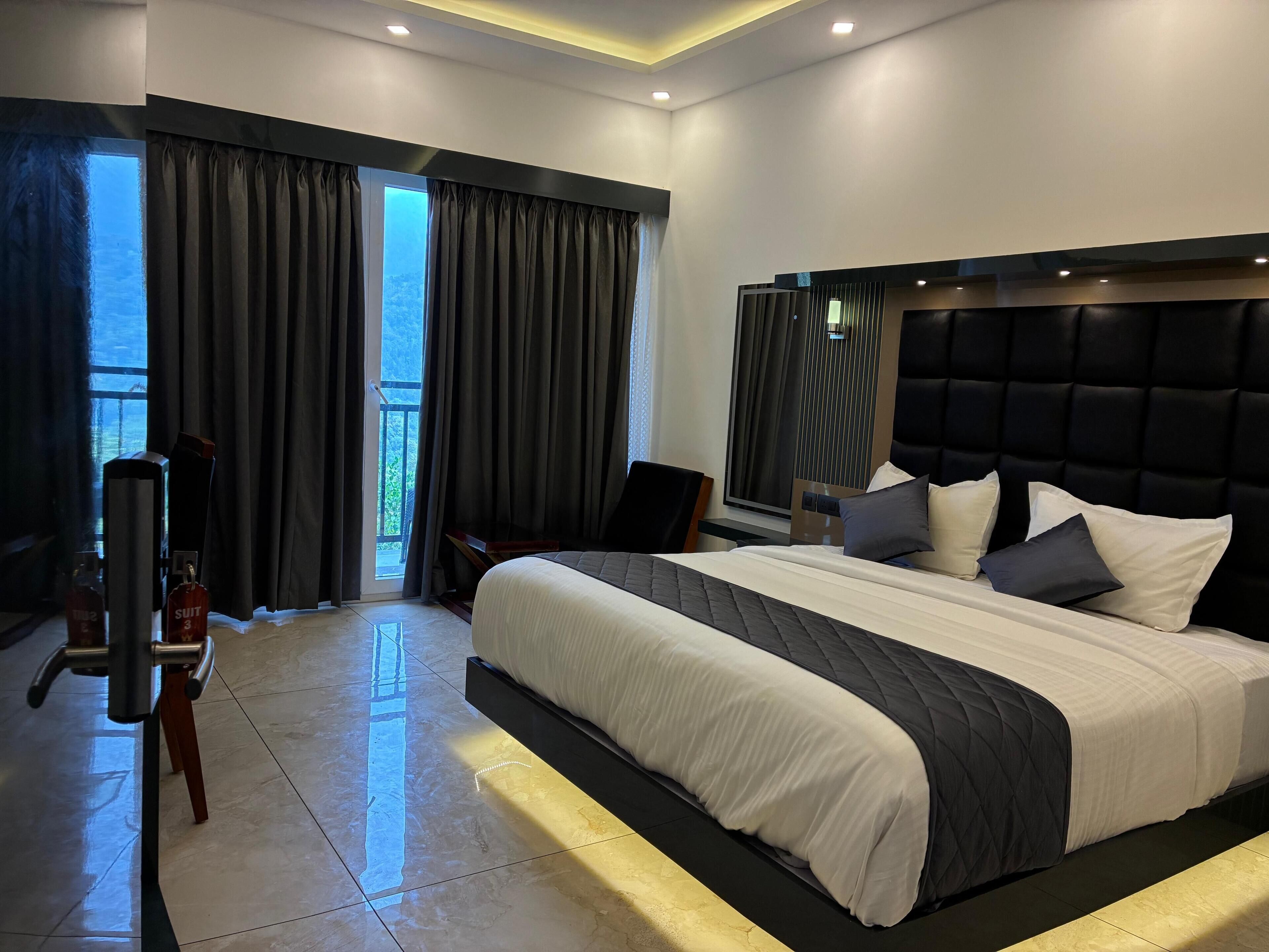 A modern hotel room with a large bed, a flat-screen TV, a seating area with a chair, and a balcony with curtains.