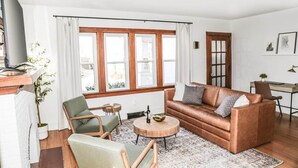 Un salon avec un canapé marron, un fauteuil vert, une table basse ronde et une grande fenêtre avec des rideaux.